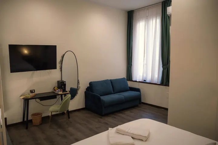 Una camera con lo scrittoio, la TV a parete, lo specchio ovale e il divano blu sotto la finestra